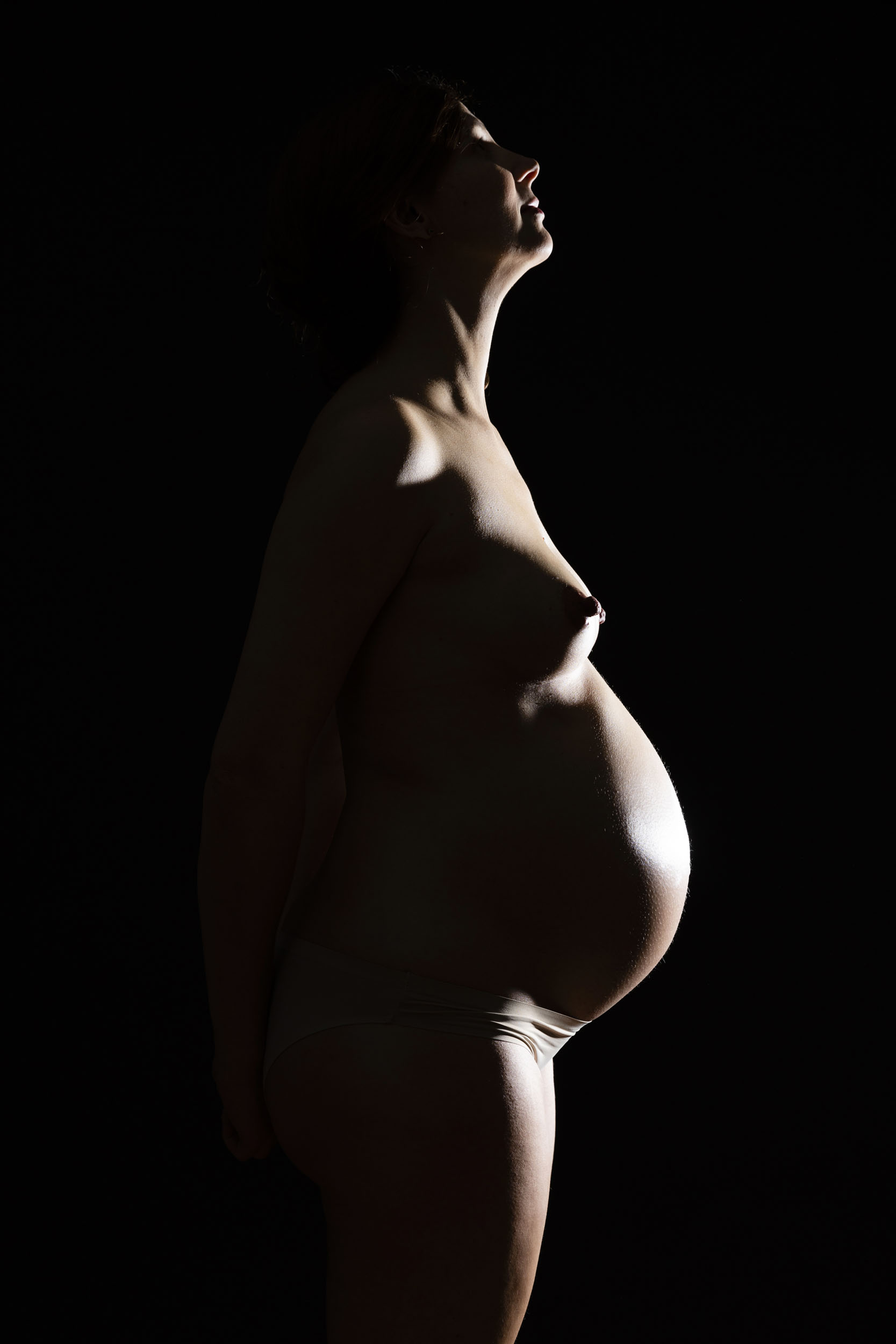 Portrait de profil d'une femme enceinte nue en contre-jour sur fond noir. Photographie de portrait réalisée en studio pour la banque d'images Plainpicture.